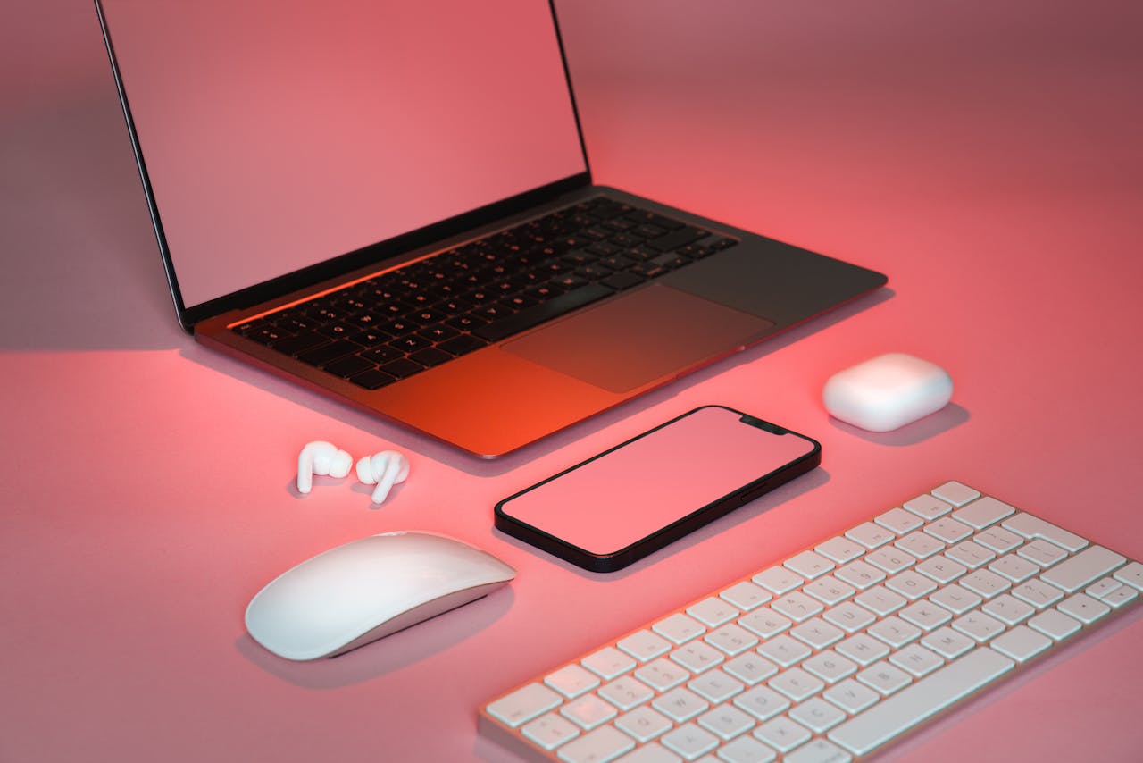 Contemporary desk setup featuring a laptop, smartphone, wireless mouse, keyboard, and earbuds in a stylish pink hue.
